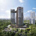 Arina East Residence. Two tall, modern towers with a connecting skybridge, surrounded by lush greenery and trees. The building features a sleek design with vertical slats and glass windows, set against a cityscape with other high-rise buildings in the background. The sky is clear with scattered clouds.