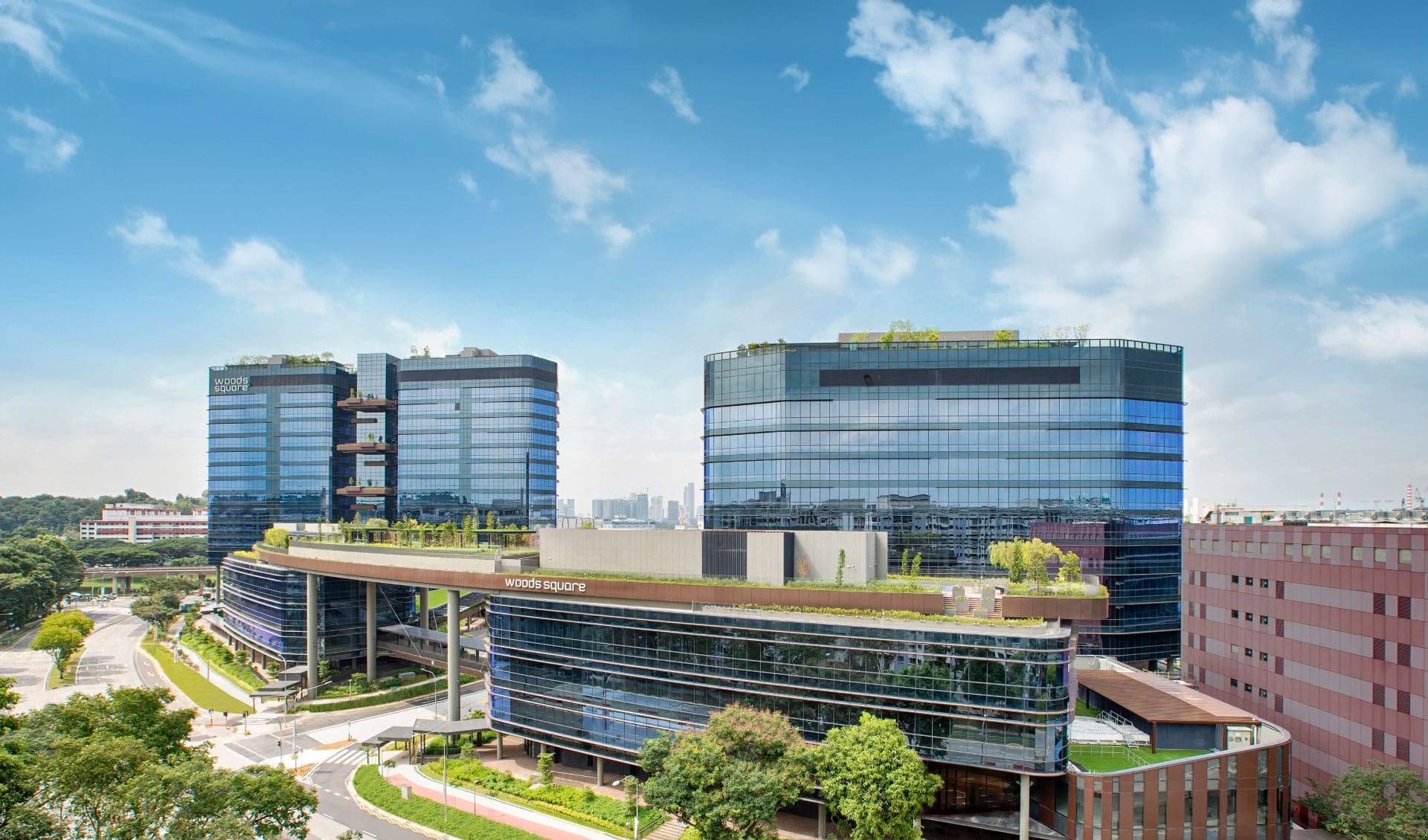 Woods Square. Modern glass towers of Woods Square in Woodlands, Singapore, with greenery on rooftops, connected by a skybridge, under a bright blue sky with scattered clouds.